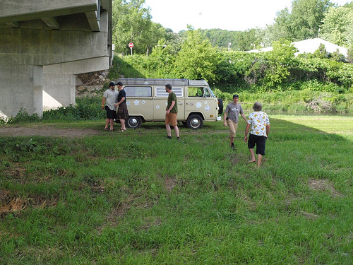 Right side view of a tan VW bus, with people standing in a grass field around, near the underside of a cement bridge